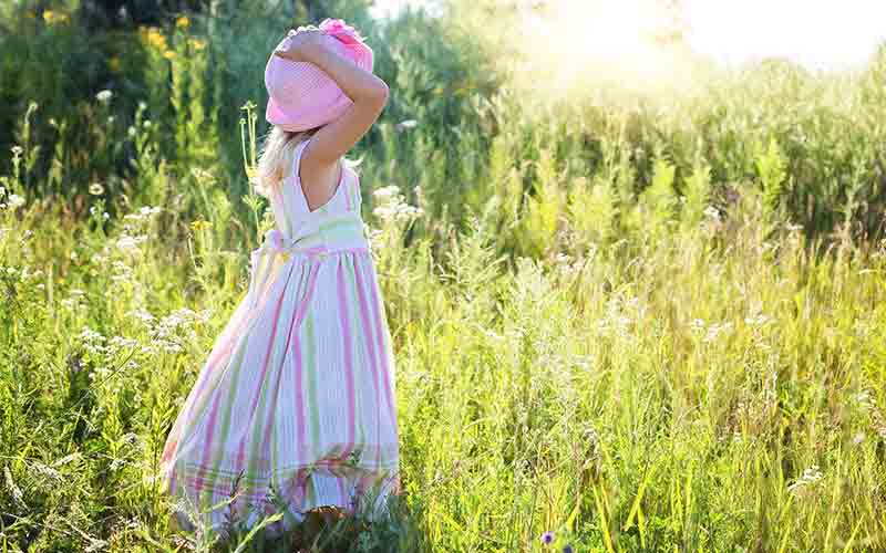 Child in a striped summer dress holding a hat while standing in sunlit tall grass